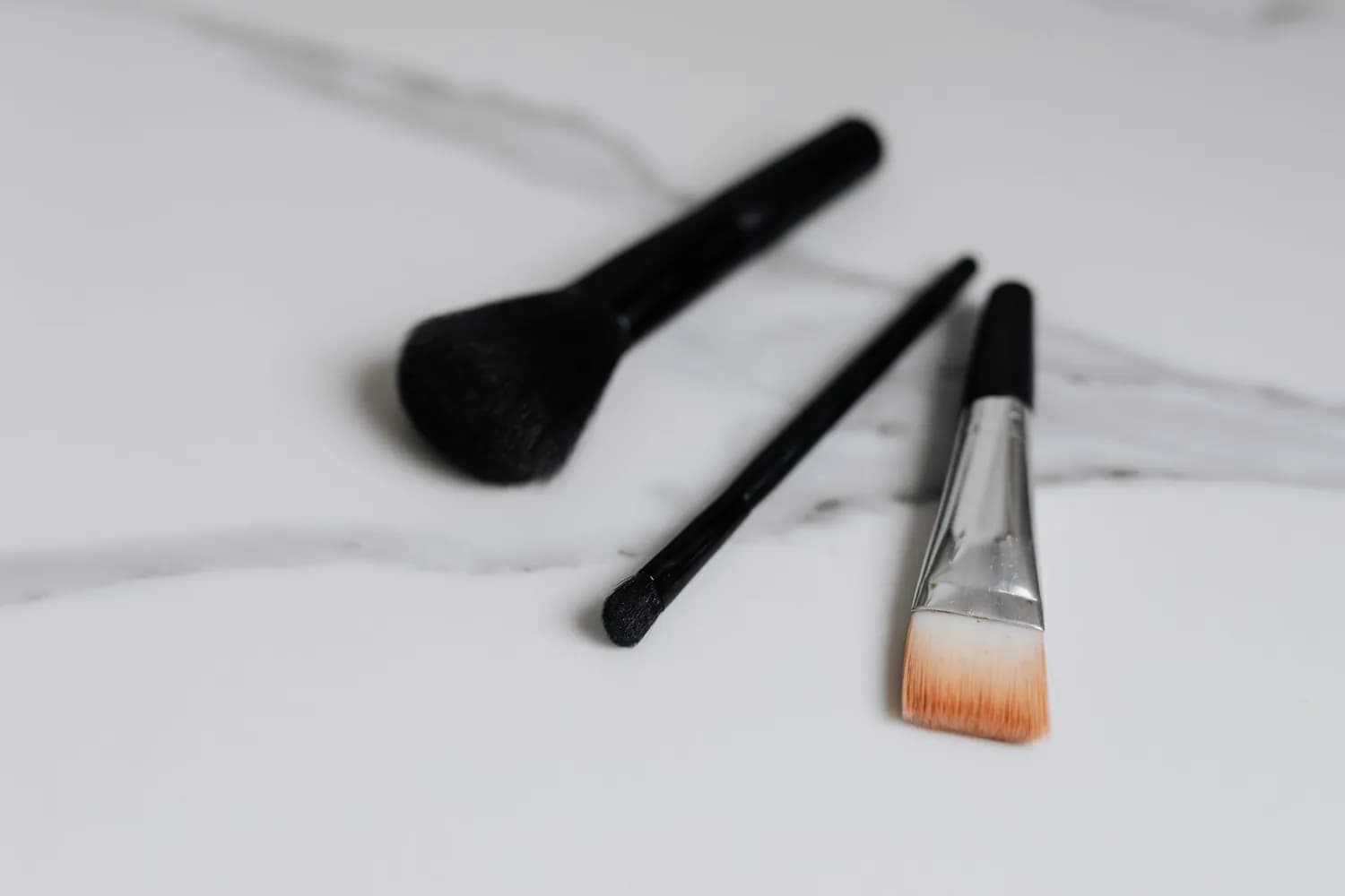 Three makeup brushes lying on a white marble surface